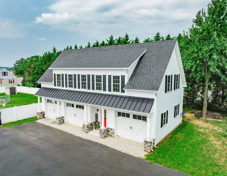 A large two-story garage with white siding, black shutters, and a black metal roof over the garage doors. The building has three garage doors framed by stone pillars at the base, and large windows on the upper level. The driveway is paved, and there is a white fence and green trees surrounding the property. The style is modern farmhouse, combining clean lines and natural materials.