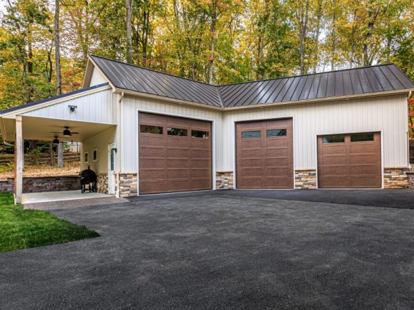 Exterior view of a three-car garage with stone accents, brown doors, and a metal roof, set against a wooded backdrop.