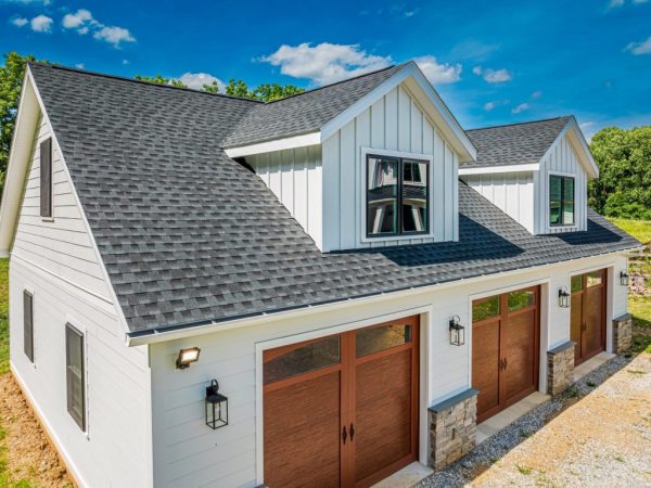 Close-up of black asphalt shingle roofing on a two-story white detached garage with dormers and wood-tone doors.
