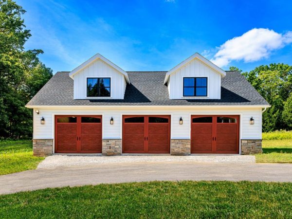 Front view of a Cape Cod-style detached garage with three wood-tone doors, stone bases, and two upper dormer windows.