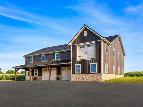 Side angle of large garage building with vertical siding, dormer-style upper door, and wide drive.