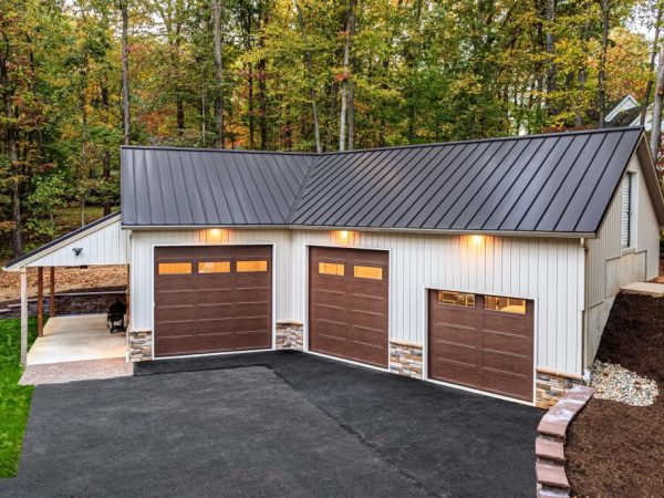 Aerial view of a three-car metal garage with stone veneer, metal roofing, and exterior lighting in a wooded area.