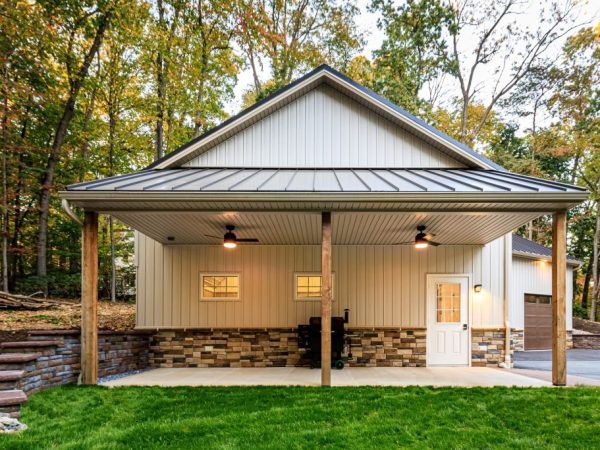 Covered side patio with ceiling fans and stone base, attached to a metal garage surrounded by grass and trees.