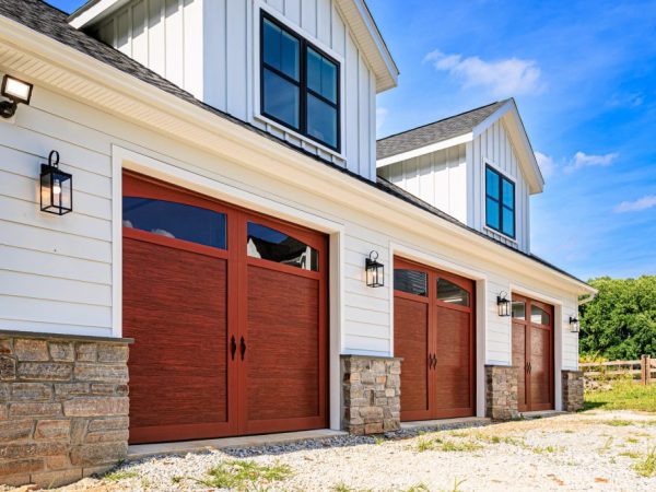 Low angle view of a white two-story garage with dark roof, faux wood doors, and a stone base trim around the perimeter.