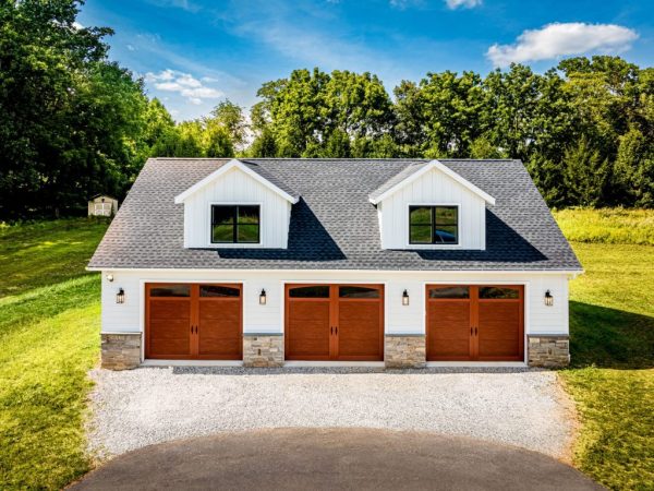 Front-facing shot of a residential three-car garage with white siding, stone pillars, and black-trimmed windows.