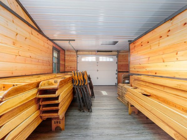 Storage room with wooden folding tables and chairs stacked along wood-paneled walls and vinyl plank flooring.