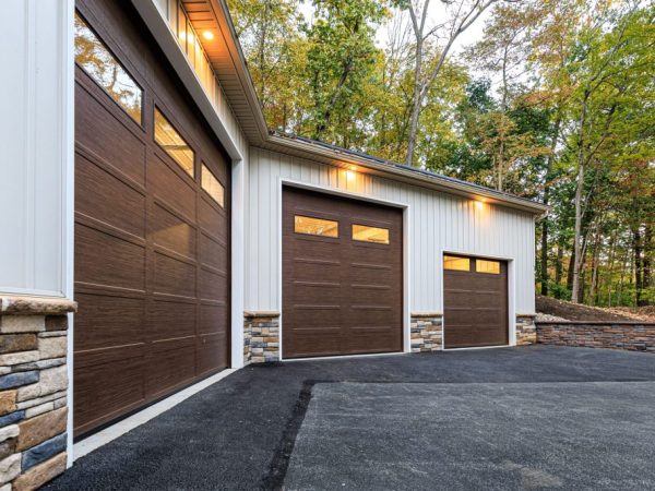 Side angle of a detached three-car garage with brown doors, stone accents, and lighting beneath a black metal roof.