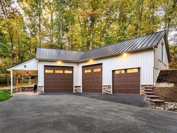 Wide shot of a detached garage with three brown overhead doors, asphalt driveway, and an attached side patio.