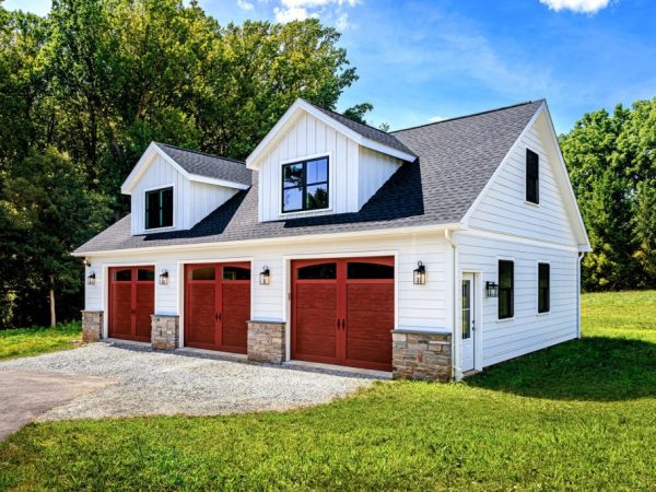 Angled view of a two-story detached garage with black roof, white siding, and three brown garage doors with upper windows.