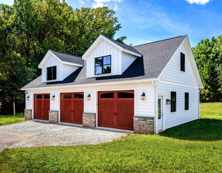 A white two-story garage with red carriage-style doors, stone accents, and a gable roof set in a grassy area surrounded by trees.