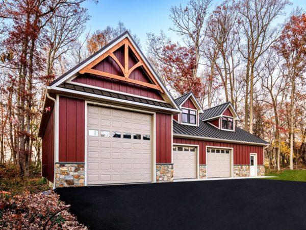 Large red detached garage with three beige overhead doors, stone wainscoting, and wood gable truss, surrounded by autumn trees.