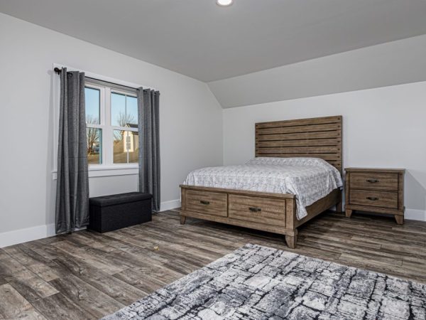 Simple bedroom setup with wooden bed, nightstand, and window with gray curtains, above a garage.