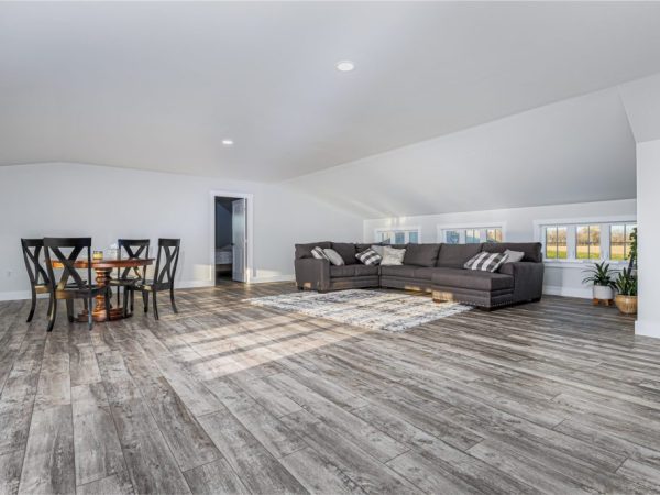 Living space with sectional sofa, dining table, and rustic flooring above a detached garage with dormer windows.