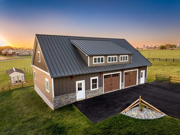 Detached garage with upper-level windows, stone base, and black metal roof, photographed at sunset in open farmland.