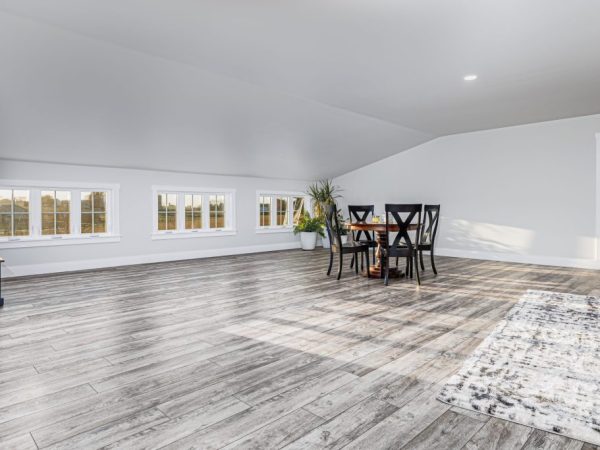 Spacious loft with gray wood flooring, multiple windows, a dining table, and sloped ceiling above a garage.
