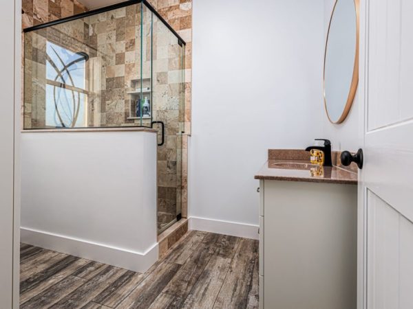 Modern bathroom with a glass-enclosed tile shower, wood-look flooring, and a single-sink vanity with round mirror.