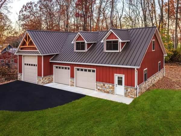 Angled view of new red garage with stone base, black metal roof, and dormers, located on a wooded lot with green lawn.