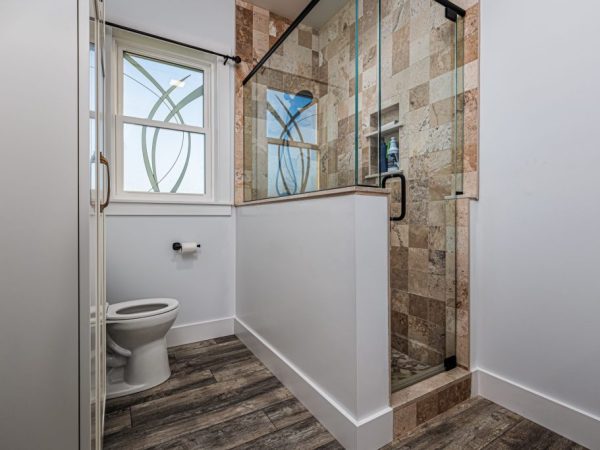Bathroom with wood tile floors, toilet, and glass-enclosed shower with decorative tile walls, located above a garage.