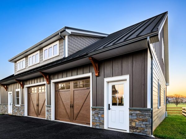 Close-up angle of two-car garage with wood-style doors, stone wainscoting, vertical siding, and black metal roof.