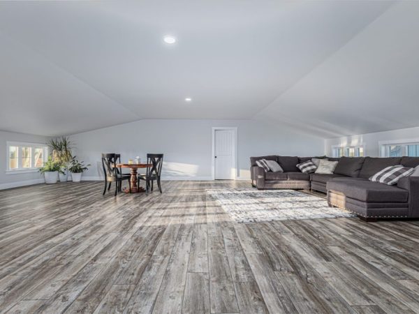 Spacious upstairs apartment above a garage featuring gray wood-look flooring, sectional sofa, and dining table.