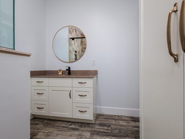 Vanity area with white cabinets, brown countertop, round mirror, and wood-look flooring in a bathroom above a garage.