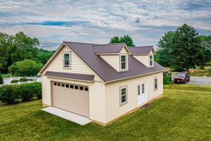 amish garage builder A beige two-story garage with dormer windows and a brown metal roof, featuring a large garage door and side entry doors, surrounded by green grass and trees.