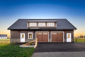 custom garage addition A dark brown custom-built garage with a black metal roof, stone accents, and a dormer window, featuring two wooden garage doors and white entry doors, set against a rural landscape at sunset.