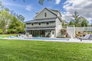 custom home addition lancaster pa A three-story gray pool house with a black metal roof, featuring large sliding glass doors, an outdoor dining area, and a swimming pool, surrounded by greenery.