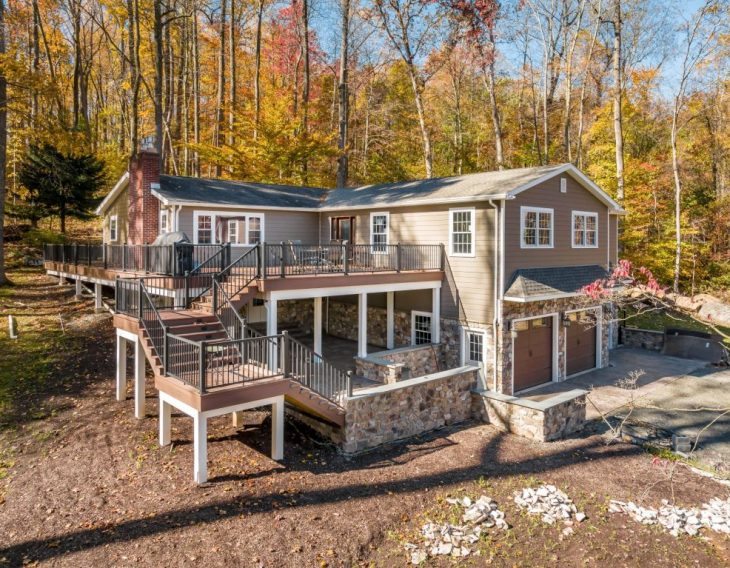 A custom-built home with a large wraparound deck, an elevated outdoor staircase, and a stone-accented lower level garage, surrounded by trees with fall colors.