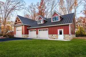 garages for sale in lancaster county A red barn-style garage with a black metal roof, stone accents, and multiple dormer windows, featuring three garage doors, including a large one for oversized vehicles, surrounded by a green lawn and autumn trees.