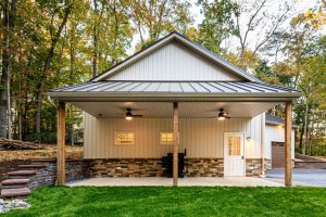 home addition company A beige detached building with a metal roof, stone foundation accents, and a covered patio area with ceiling fans, set against a wooded backdrop.