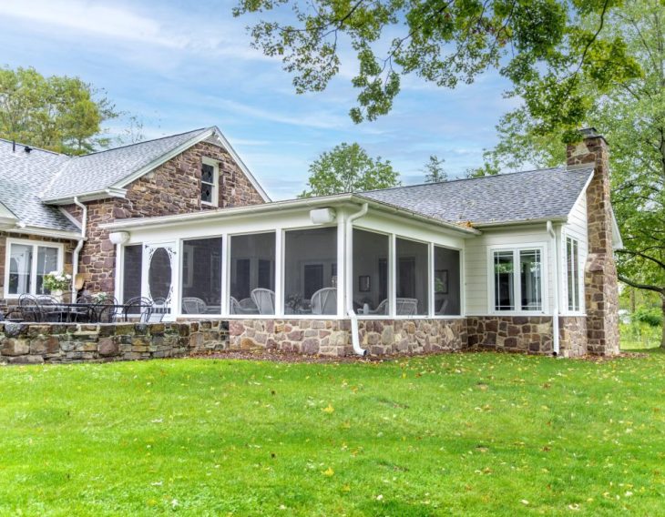 A stone farmhouse with a screened-in porch addition, featuring white trim and a shingled roof, surrounded by a green lawn.