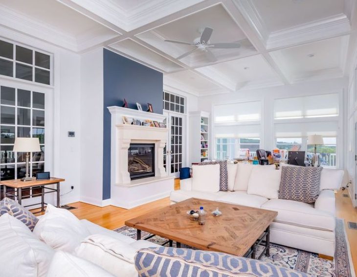 A modern living room with white coffered ceilings, a built-in fireplace with a blue accent wall, large windows, and a white sectional sofa, featuring a wooden coffee table and decorative pillows.