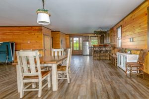 A rustic open-concept dining and kitchen area with wooden walls and flooring, featuring a farmhouse-style dining table, a bar with wooden stools, and a glass door leading outside.