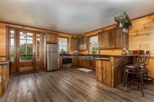  A rustic kitchen with wood-paneled walls, dark-stained wooden cabinets, a farmhouse sink, and a stainless steel refrigerator, featuring a breakfast bar with barstools and glass doors leading outside.