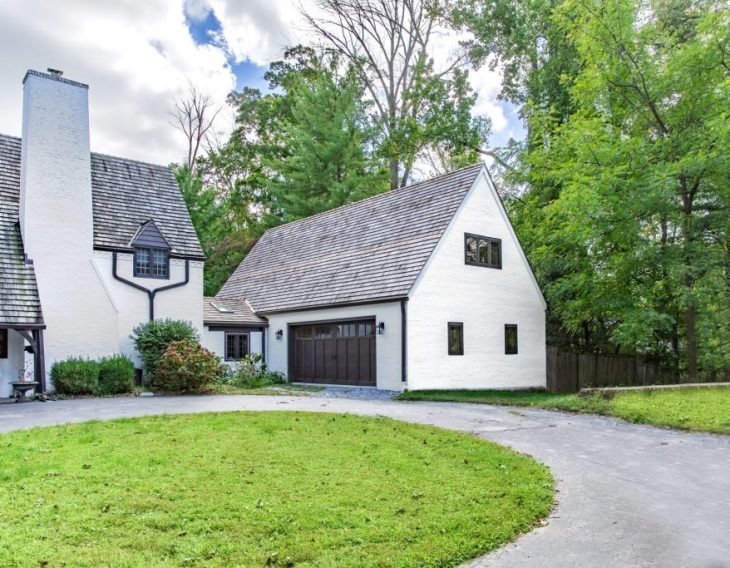 A Tudor-style home with a white brick exterior, steeply pitched roof, and dark trim, featuring a detached garage with a matching design, surrounded by trees and a curved driveway.