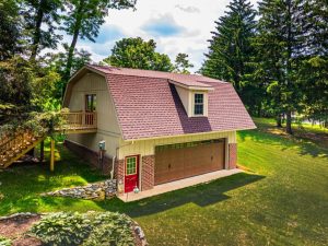 lancaster home addition A beige barn-style garage with a brown shingle roof, dormer window, brick lower walls, and a wooden garage door, featuring an exterior staircase leading to a second-floor entrance, set in a tree-filled yard.