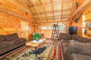 A cozy cabin-style living space with exposed wood beams, log walls, and a vaulted ceiling, furnished with brown sofas, a live-edge coffee table, and a decorative ladder-style shelf.