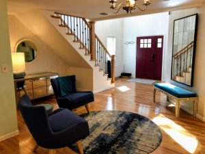 A modern entryway with a red front door, hardwood floors, a staircase with wrought iron railings, and a seating area with blue chairs, a round rug, and a mirrored accent table.