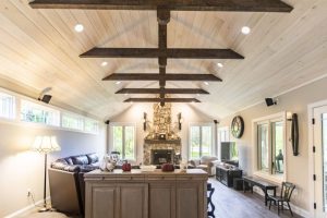 A vaulted-ceiling living room with exposed wood beams, a stone fireplace, and large windows, featuring a mix of modern and rustic furnishings, including a leather sofa and a farmhouse-style island.