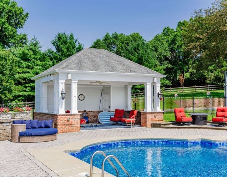 A poolside cabana with white columns, a shingled roof, and brick accents, featuring outdoor seating and a ceiling fan, next to a blue in-ground pool.