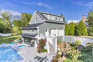 A modern three-story pool house with a gray exterior, black metal roof, and an upper-level garage, featuring a wraparound patio and stairs leading to a landscaped pool area.