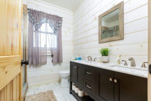 A farmhouse-style bathroom with white shiplap walls, a dark double vanity with a marble countertop, a framed mirror, and a glass-enclosed shower, illuminated by natural light from a window with sheer curtains.