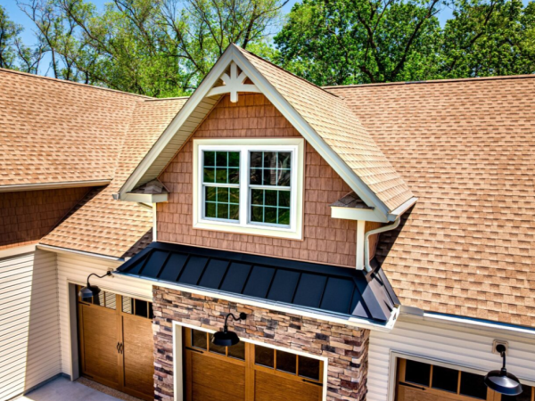 close up of a dormer window above three garage doors, featuring brown shake siding, and a black metal awning